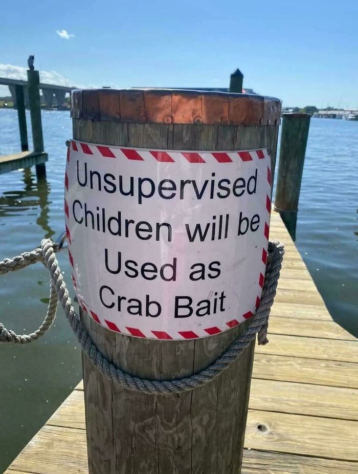 Sign on dock pole humorously warning that unsupervised children will be used as crab bait, a blursed image near water.