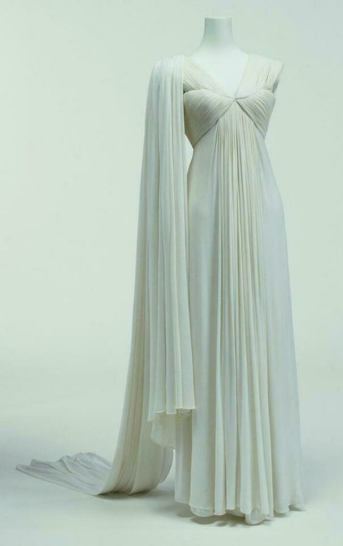 White flowing gown with pleated fabric and draped sleeve, showcasing centuries of fashion on a mannequin in a studio setting.