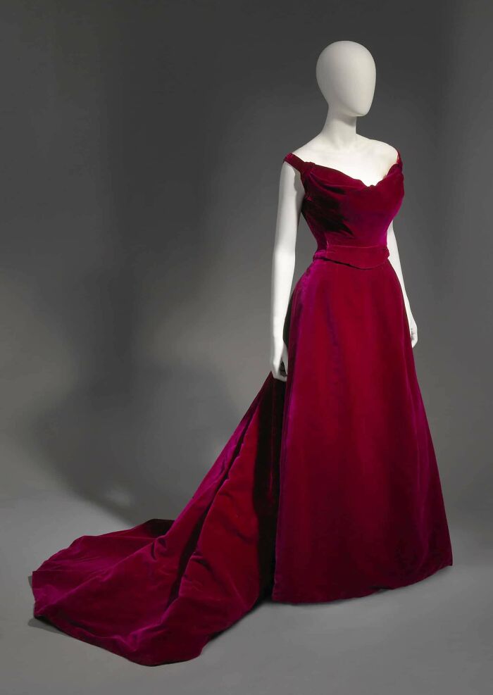 Mannequin wearing a deep red velvet gown with a long train, showcasing centuries of fashion in a museum setting.