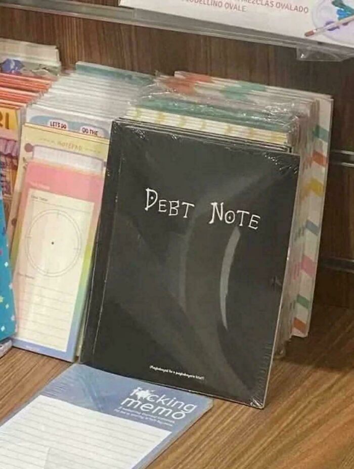 Black notebook labeled Debt Note standing among colorful notepads in a display, a blursed image causing confusion.