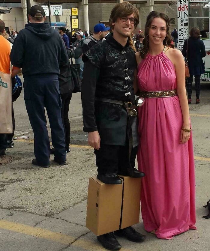 A blursed image of a man standing on a box that aligns with his shoes, next to a woman in a pink dress at an outdoor event.