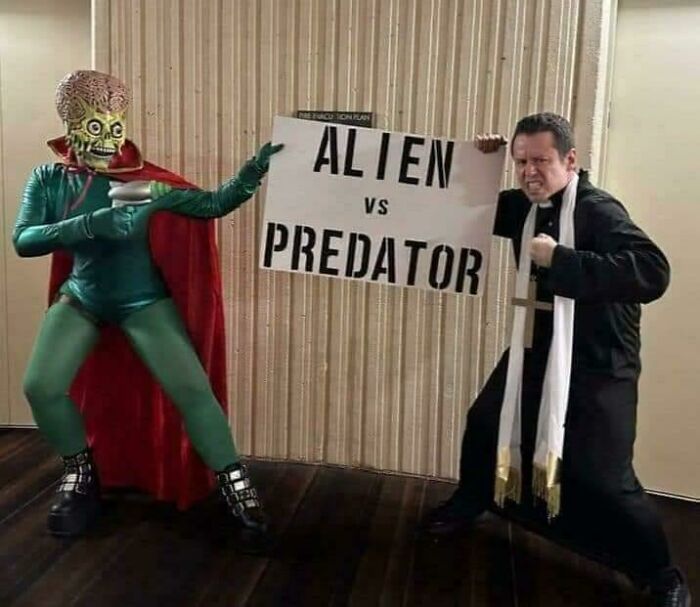Person in alien costume facing man dressed as priest holding alien vs predator sign in blursed image with surreal vibe