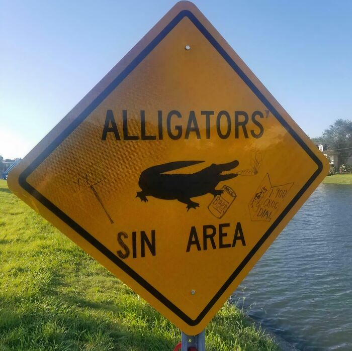 Yellow street sign humorously altered by wholesome vandals showing an alligator in a sin area near a water body.