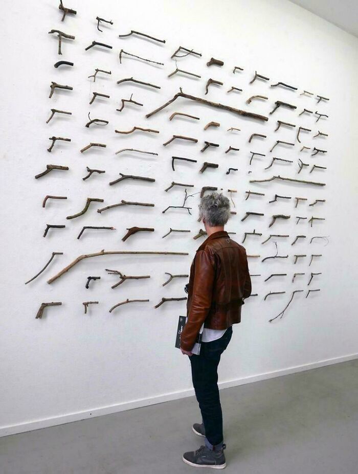 Person in a brown jacket observing an art installation of various oddly shaped sticks on a white wall, blursed images concept.