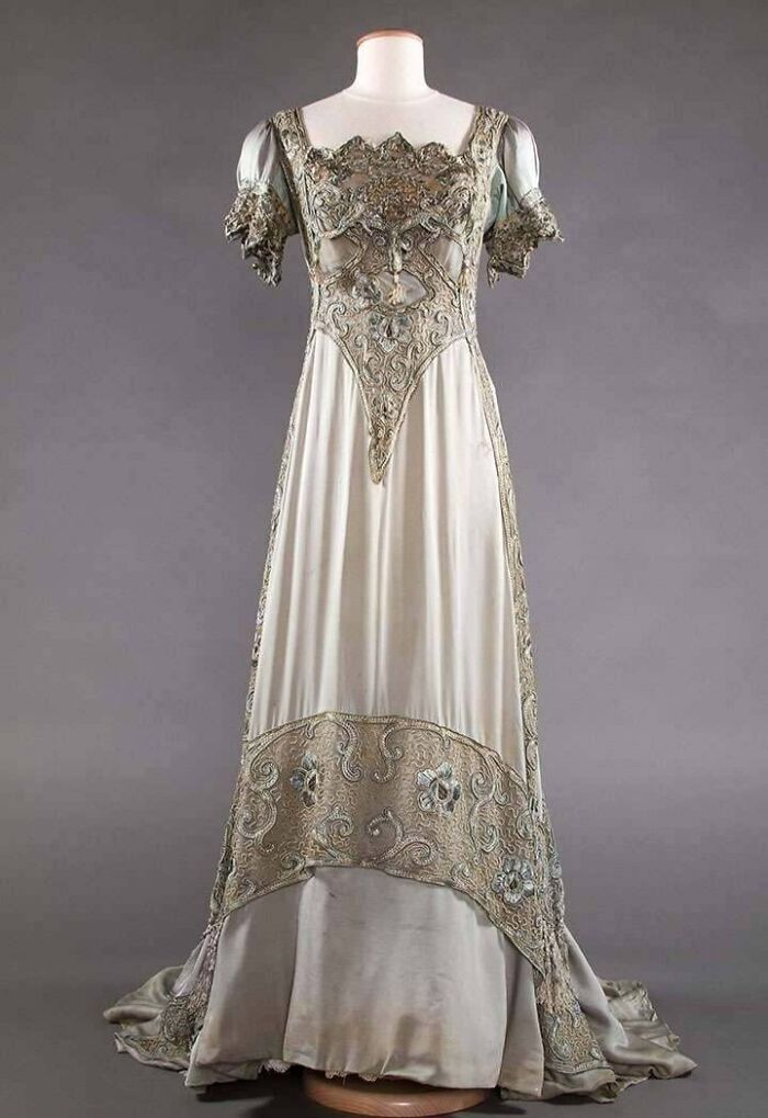 Elegant vintage dress with intricate embroidery showcasing centuries of fashion history on a plain gray background.