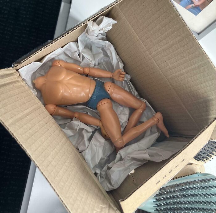 A creepy headless male action figure in blue underwear lying in a cardboard box with crumpled paper packaging.