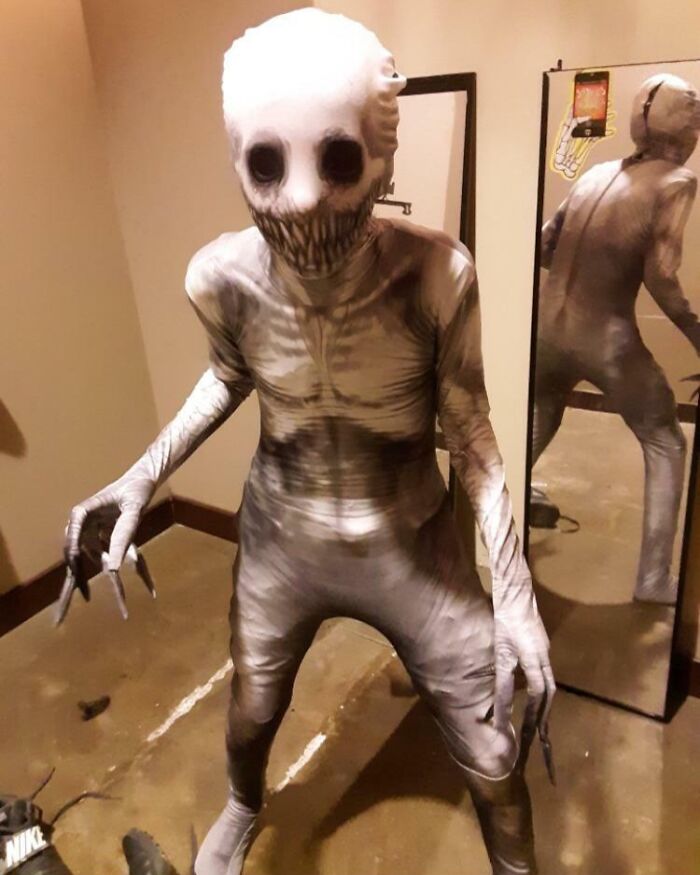 Creepy figure in a tight, skull-like costume with dark eyes and sharp teeth standing in a dim room near a mirror.