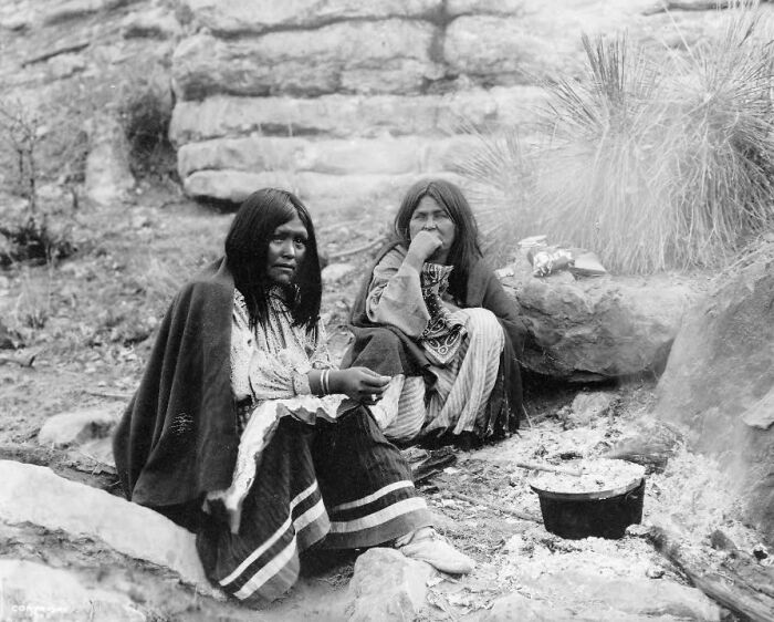 Two Native American women in traditional clothing sitting outdoors by a fire, revealing Native American life in the 1900s.