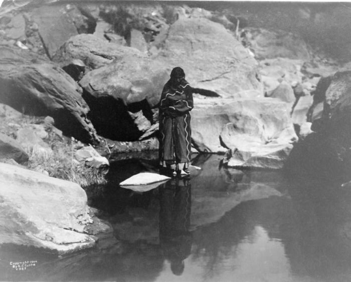 Native American woman in traditional clothing standing by a rocky stream reflecting 1900s life.