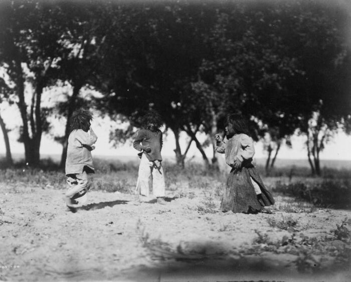 Three Native American children playing outdoors in the early 1900s, surrounded by trees and natural landscape.