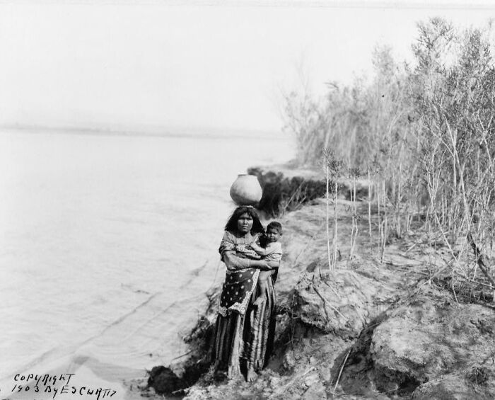 Native American woman carrying a pot on her head while holding a child near a riverbank in the early 1900s.