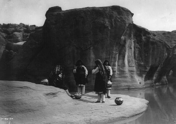 Native American life in the 1900s shown with women gathering by rocky riverbank in traditional clothing.