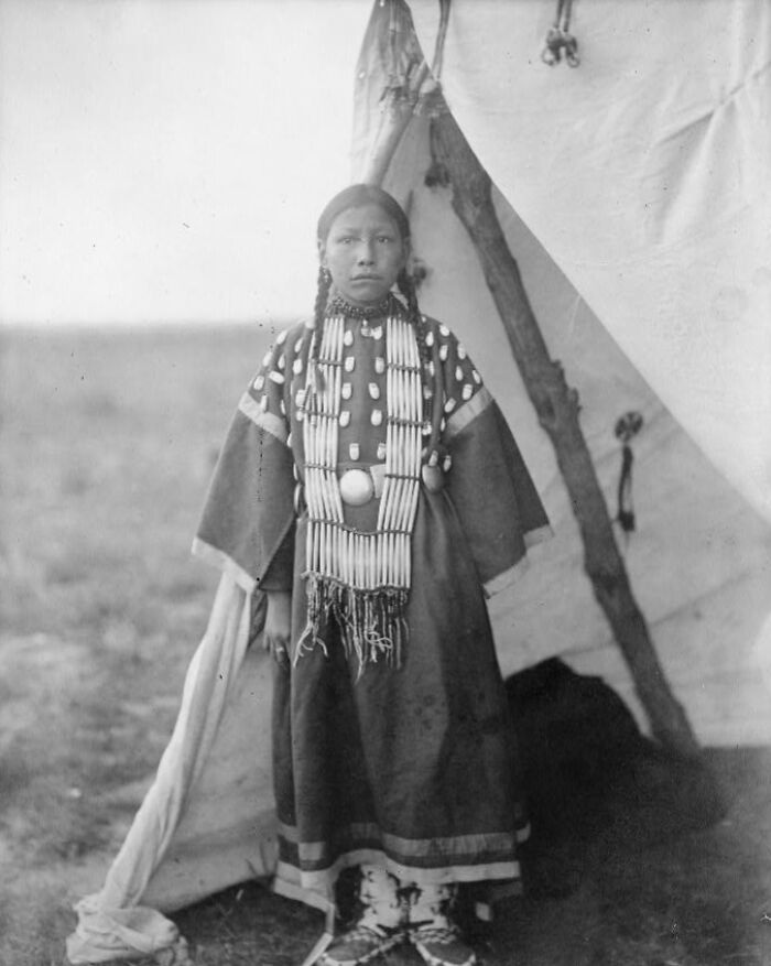 Native American girl dressed in traditional clothing standing outside a teepee, showcasing Native American life in the 1900s.