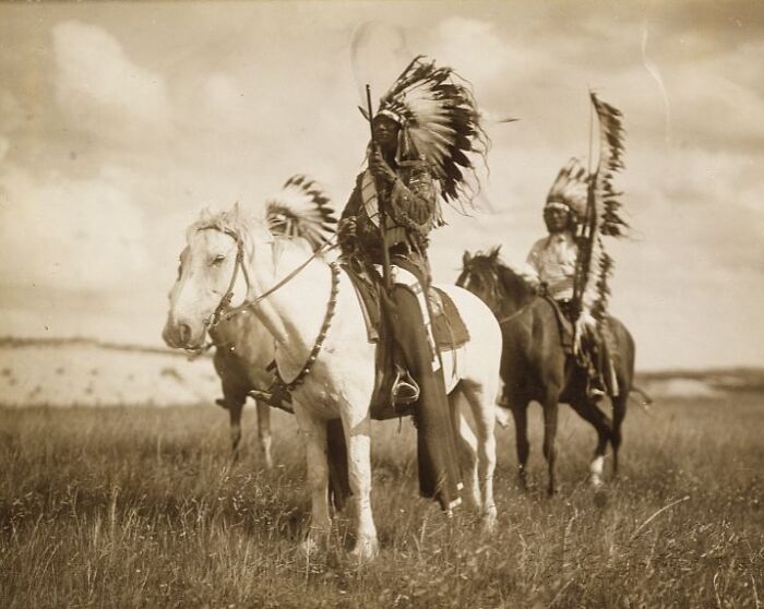 Native American men in traditional headdresses riding horses on open plains, showcasing life in the 1900s.