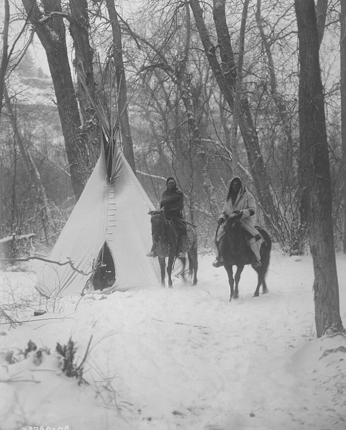 Two Native Americans on horseback near a teepee in a snowy forest, illustrating Native American life in the 1900s.
