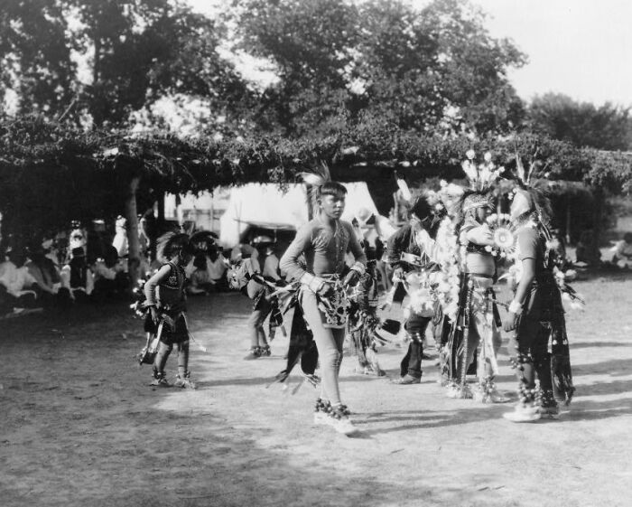 Native Americans in traditional attire performing a dance in a 1900s outdoor cultural gathering.
