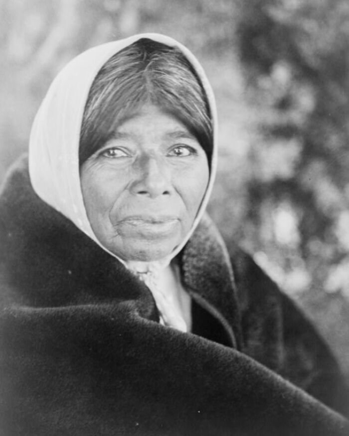 Native American woman from the 1900s wrapped in a blanket and wearing a headscarf, showing life in that era.
