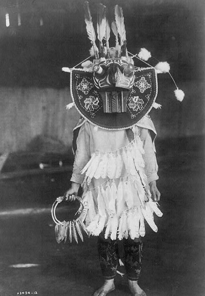Native American wearing traditional ceremonial costume with feathered mask and holding decorated hoop in the 1900s.