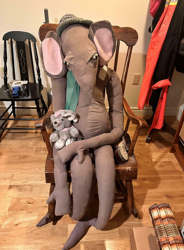 Large creepy elephant doll with mismatched eyes holding a small plush bunny, seated in a wooden chair indoors.