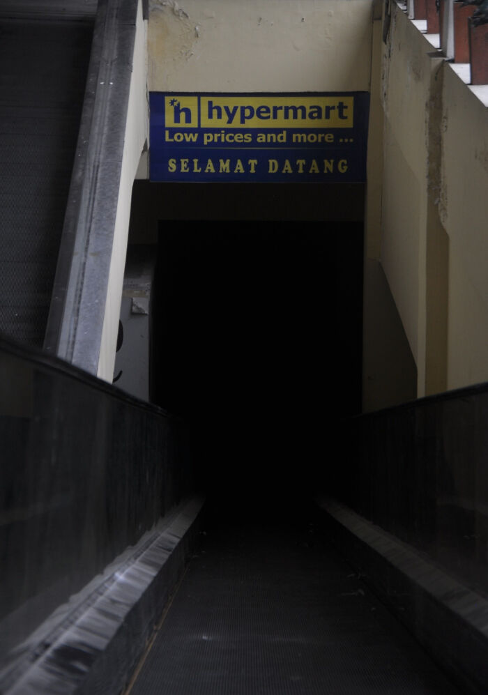 Dark, creepy escalator entrance beneath a worn sign for hypermart, evoking an eerie and unsettling atmosphere.