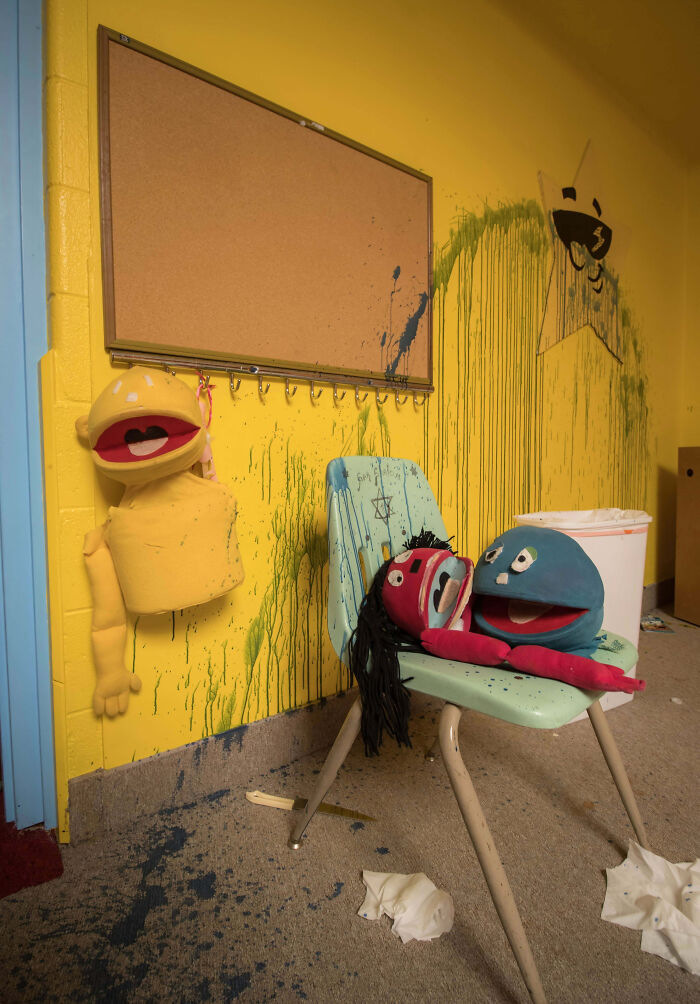 Creepy puppet heads and splattered paint in a worn yellow room with a bulletin board and scattered trash on the floor.