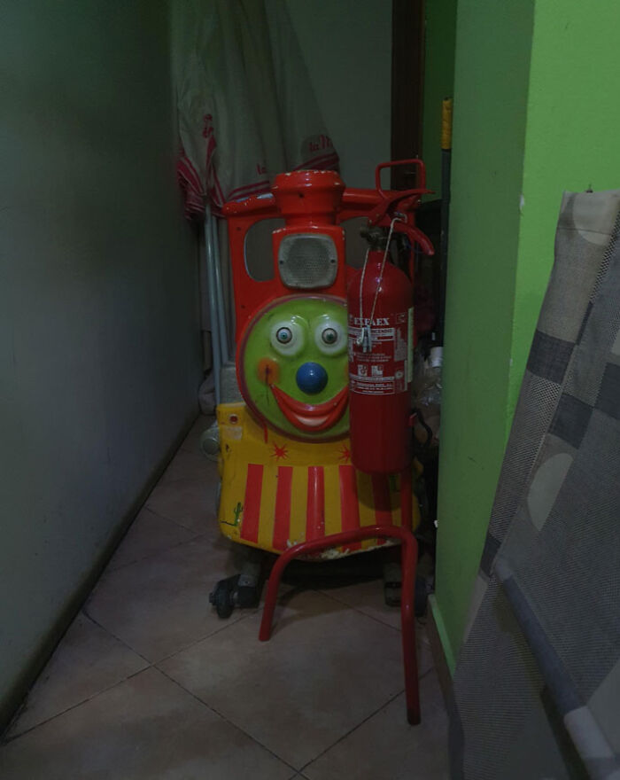 Creepy clown-faced train toy hidden in a dim hallway next to a fire extinguisher and household items.