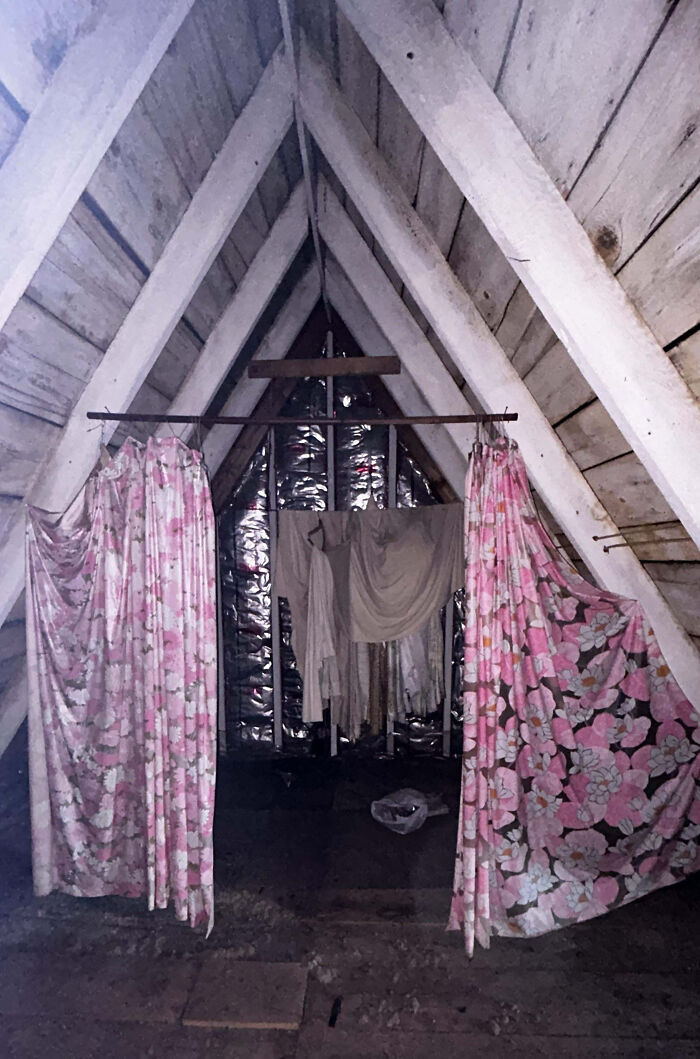 Creepy attic with aged wooden beams, hanging floral curtains, and old clothes draped in dim light.