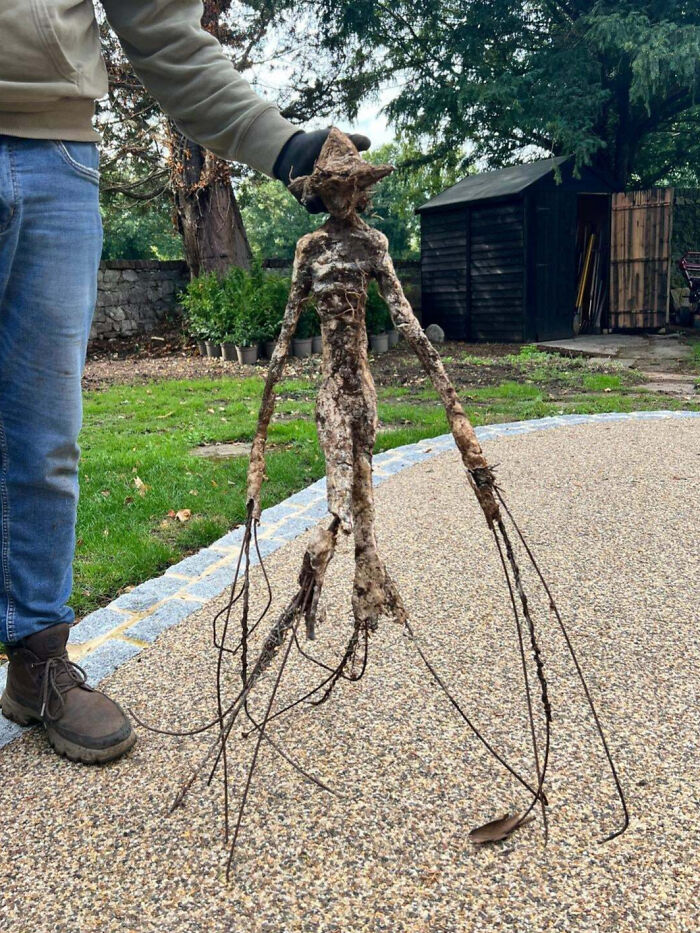 Person holding creepy root shaped like a humanoid figure with long wire-like extensions outdoors on a stone path.