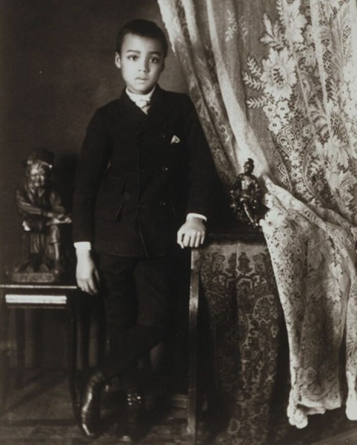 Young African American boy in a suit posing indoors, captured in early 1900s photo highlighting African American life.
