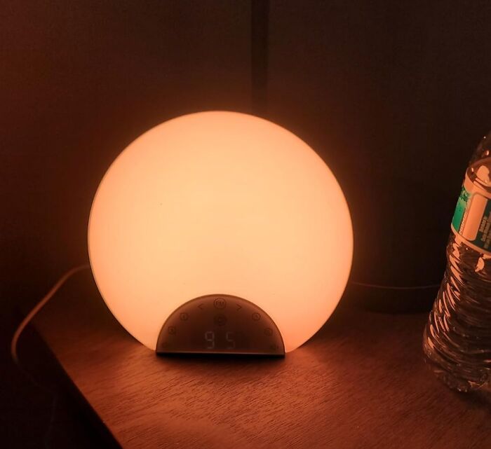 Sunrise alarm clock emitting warm light on wooden nightstand, helping to hack your way into becoming a morning person.