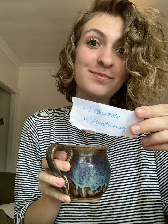 Young woman holding a ceramic mug and a note while participating in an internet toast challenge with kind comments.