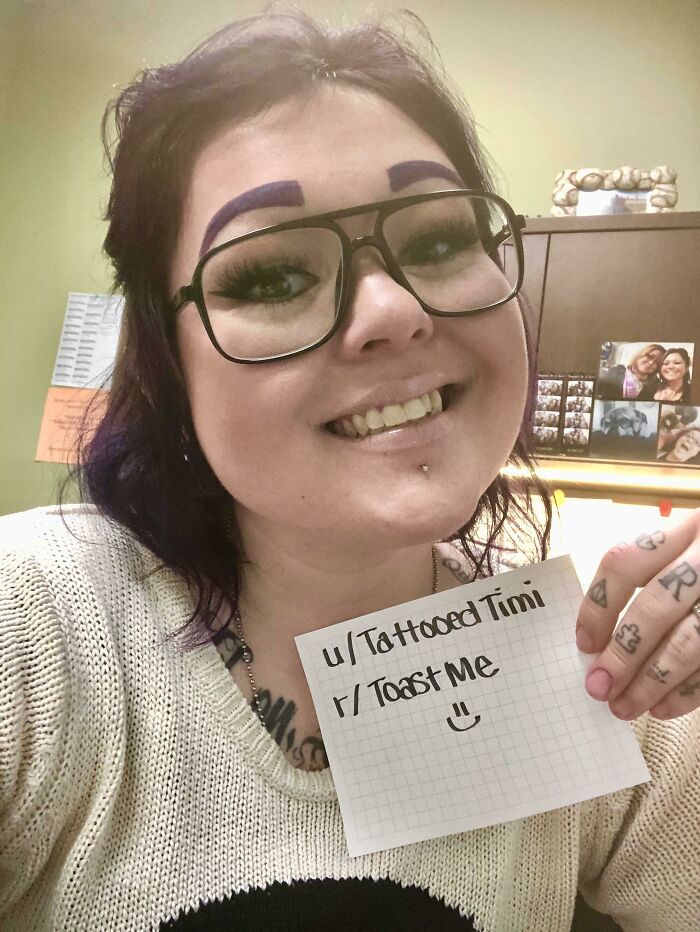 Person holding a toast me sign with tattoos and purple eyebrows, smiling in a casual indoor setting for internet toast comments.