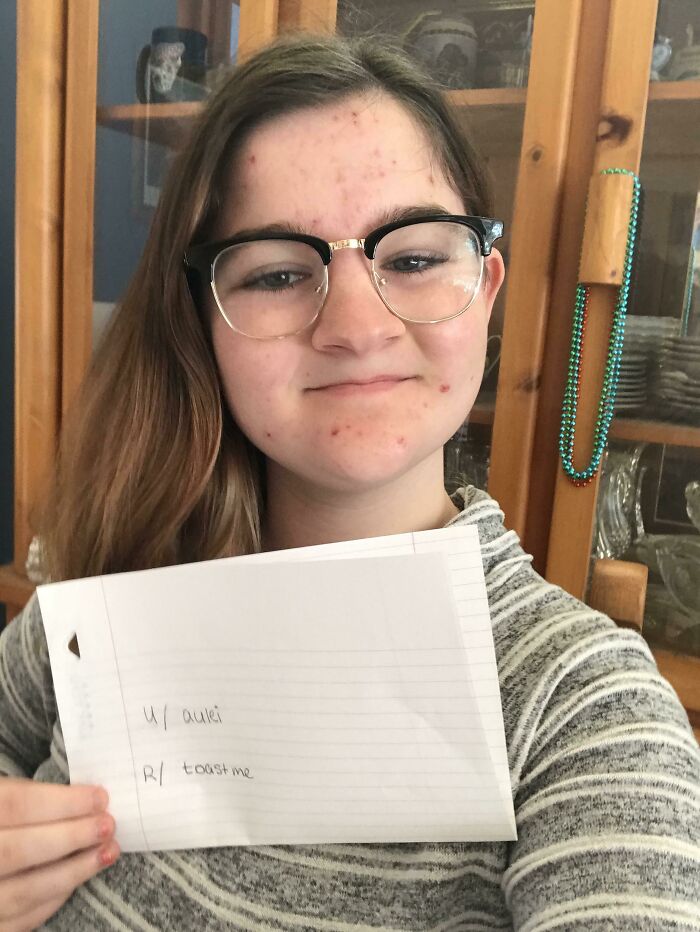 Young woman holding a note for a toast me request, smiling gently in a cozy home setting with glasses and casual wear.