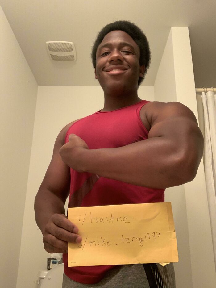 Smiling muscular man in a red tank top flexing arm while holding toast request paper in a bathroom setting.