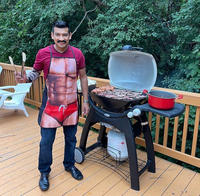 Man wearing chaotic novelty apron grilling meat on a backyard deck with trees in the background.