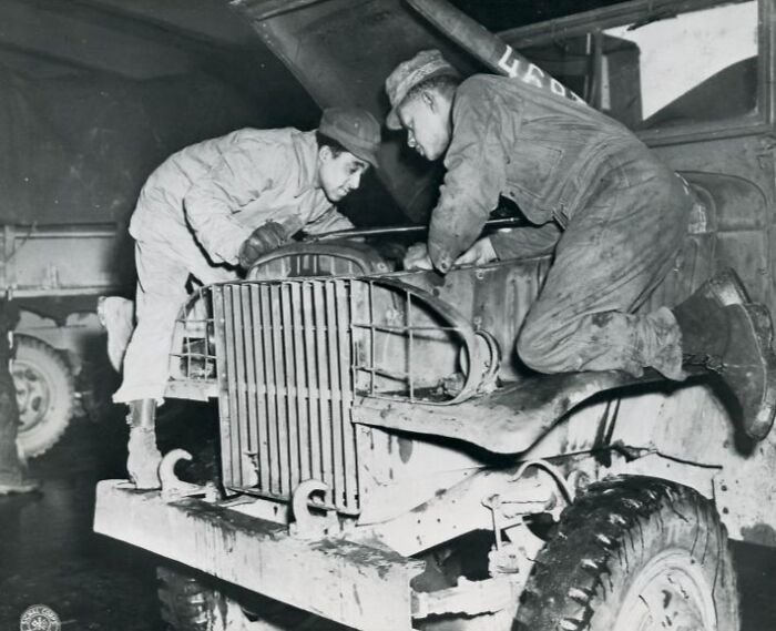 Two African American soldiers in uniform repairing a military vehicle inside a garage during wartime.