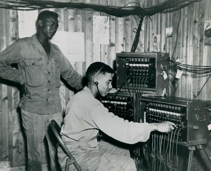 Two African American men in military uniform operating vintage communication equipment in a wood-paneled room.