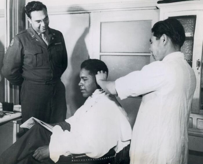 African American soldier in uniform getting a haircut while another uniformed man watches in a historic setting.