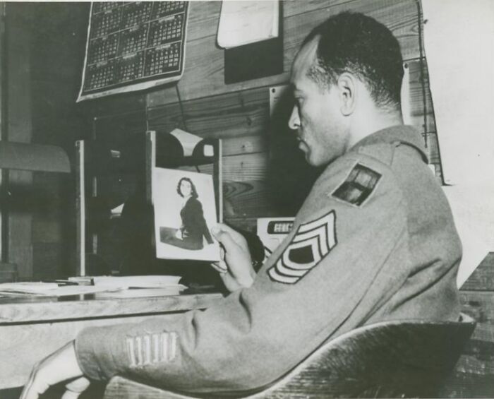 African American soldier in uniform sitting at desk looking at a photo in a historical military setting.