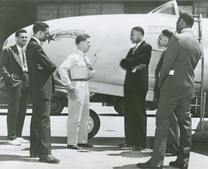 Group of African Americans in uniform and suits standing near a military aircraft during a historical moment in aviation.