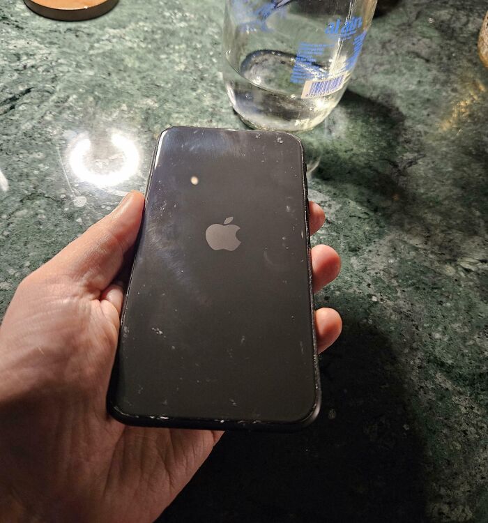 Hand holding a scratched Apple smartphone showing a small mark that resembles a one-in-a-million coincidence on the screen.