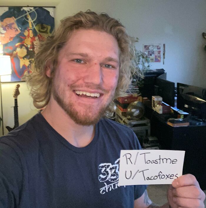 Young man holding a Toast request sign, smiling in a room filled with gaming and music posters for positive internet comments.