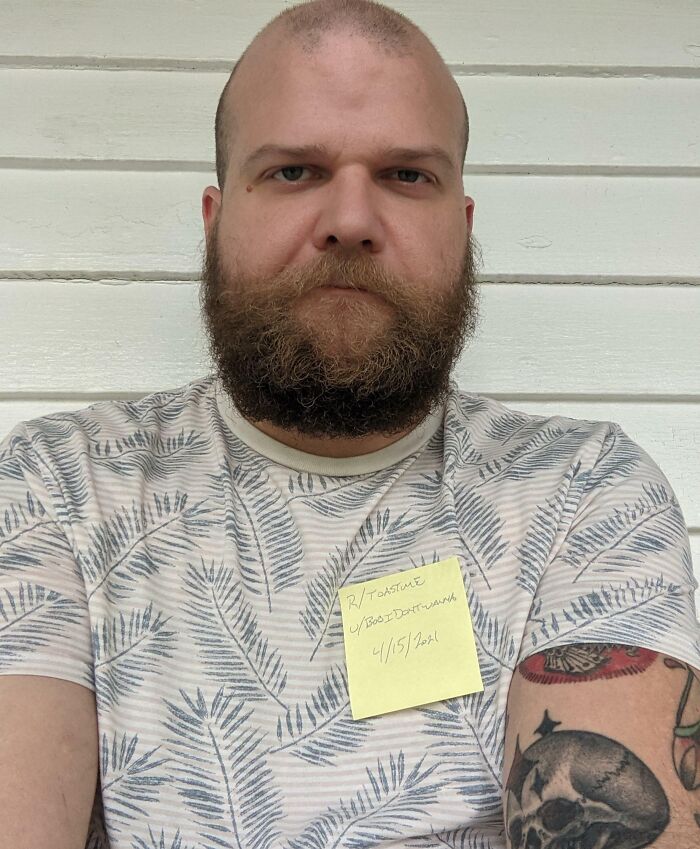 Bearded man with a tattoo wearing a patterned shirt and holding a note for the internet toast challenge.
