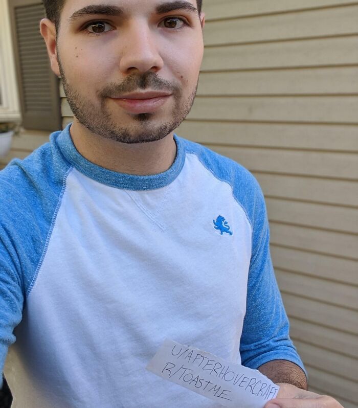 Young man holding a note requesting a toast, smiling in a casual blue and white shirt outside a house.