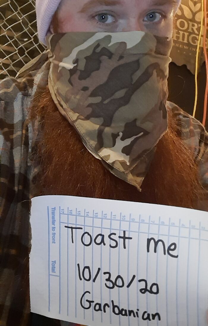 Person with a red beard wearing a camo face covering holding a sign asking the internet to toast them for kind comments.