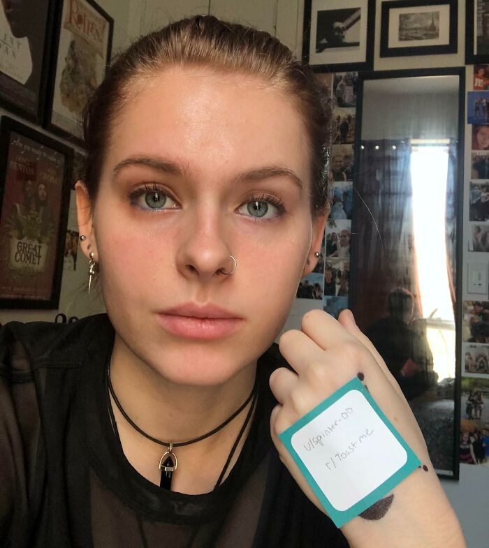 Young person holding note for internet toast, smiling softly with nose ring and piercings in casual room setting.