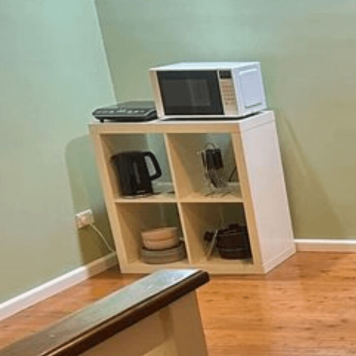 Microwave and kitchen appliances poorly placed on a small shelf in a corner of a rented room showing bad landlord setup.