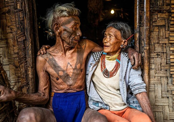 Elderly couple with traditional tattoos and jewelry sitting closely in a rustic setting, captured in striking portrait style