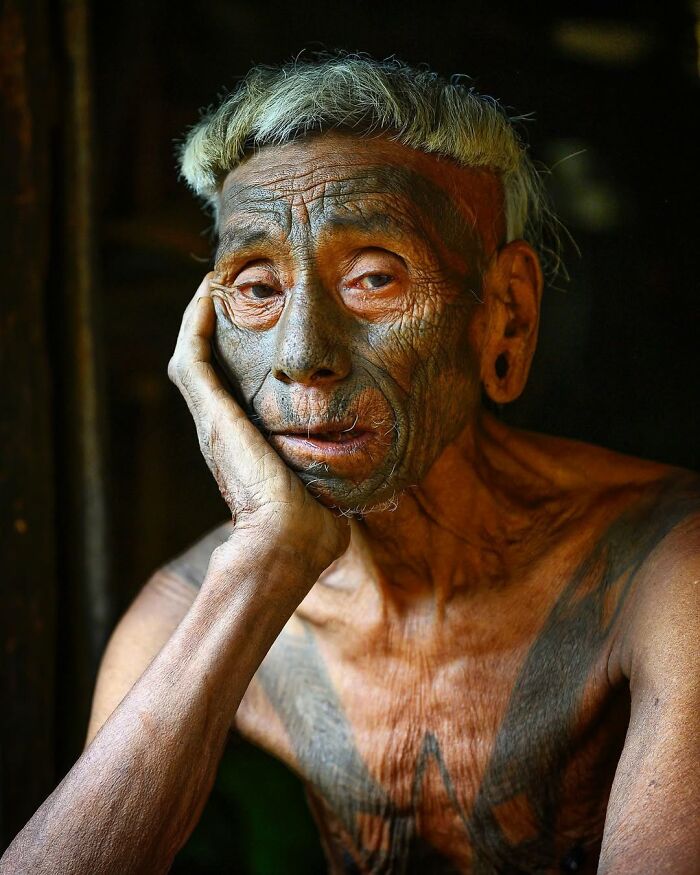 Elderly person with traditional facial tattoos resting head on hand in a striking portrait by Massimo Bietti.