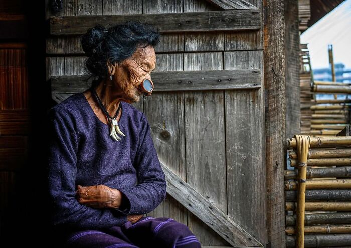 Elderly woman with traditional nose ornament sitting inside rustic wooden house, captured in striking portrait across the globe.